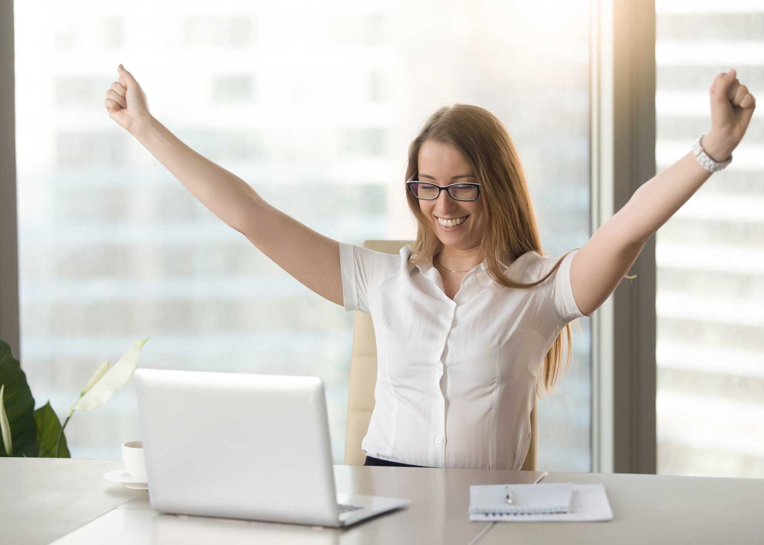 A young woman sitting at a desk in front of a window looking at a white laptop throws her arms up on a gesture of victory.